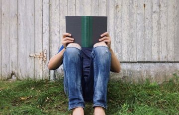 Bare-foot man in jeans sitting outside on grass reading 'American Phinx', his face is covered by the book